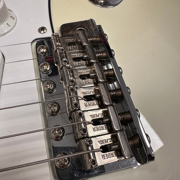 Close-up of Fender guitar chrome bridge saddle with six adjustable string saddles and white guitar body.