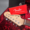 Close-up of Fender Stratocaster electric guitar headstock with tuning pegs, resting in red plush guitar case with red Fender registration card and black leather case accessory.