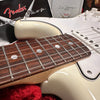 Close-up of Fender Stratocaster electric guitar rosewood fretboard and white body with vintage-style black and white binding, showing strings and part of the pickguard and controls, with Fender logo and documentation in the background.