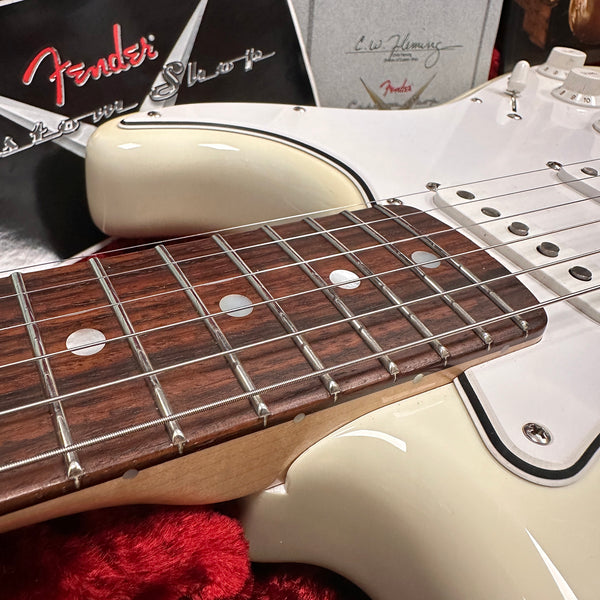 Close-up of Fender Stratocaster electric guitar rosewood fretboard and white body with vintage-style black and white binding, showing strings and part of the pickguard and controls, with Fender logo and documentation in the background.