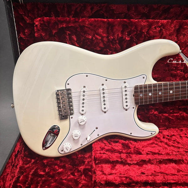 Close-up of white electric guitar body with three single-coil pickups and white pickguard inside a red plush-lined guitar case