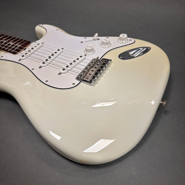Close-up of white electric guitar body with white pickguard, three single-coil pickups, volume and tone control knobs, and chrome bridge on gray background