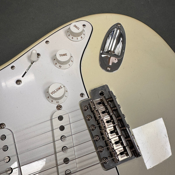 Close-up of white electric guitar body showing tone and volume knobs, pickup selector switch, chrome bridge with Fender branding, and cream pickguard.