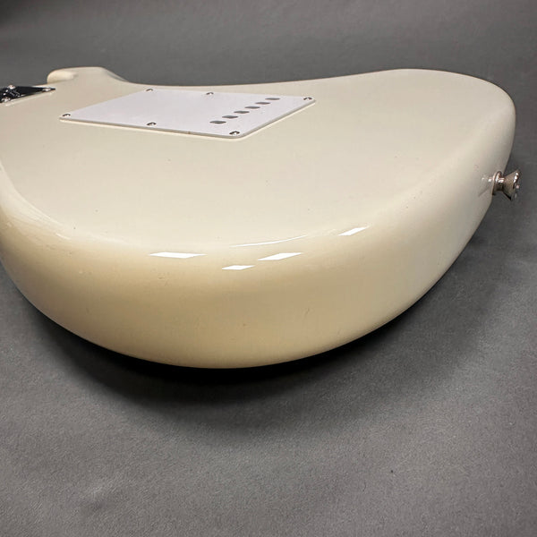 Close-up view of the back side of a white electric guitar body with a white plastic tremolo cavity cover and strap button on a gray surface