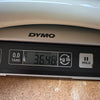 Close-up of a Dymo digital scale displaying a weight of 3.646 kilograms on a wooden surface, used for weighing guitars or guitar accessories.