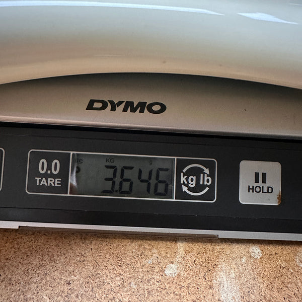 Close-up of a Dymo digital scale displaying a weight of 3.646 kilograms on a wooden surface, used for weighing guitars or guitar accessories.