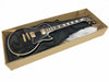 Black electric guitar with gold hardware and white binding inside a cardboard shipping box with a black gig bag underneath