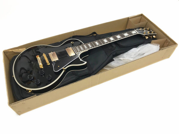 Black electric guitar with gold hardware and white binding inside a cardboard shipping box with a black gig bag underneath