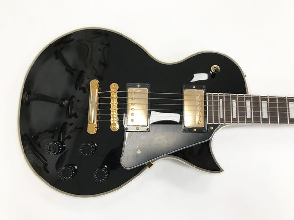 Black electric guitar with gold hardware, dual humbucker pickups, black pickguard, and four control knobs on white background