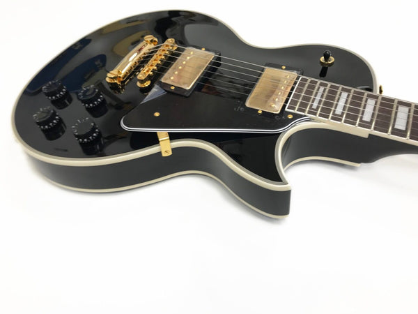 Close-up of black electric guitar with gold hardware, dual humbucker pickups, black control knobs, and cream binding on body and neck against white background.
