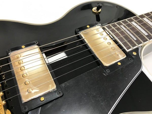 Close-up of black electric guitar body showing gold humbucker pickups, strings, and pickup selector switch with white binding around edges