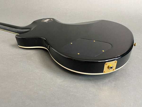 Black electric guitar body back with gold input jack and cream binding edge on gray background