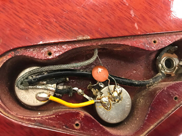 Close-up view of vintage electric guitar control cavity showing potentiometers, capacitor, and wiring inside red wooden body.