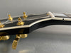 Close-up of black electric guitar headstock with gold tuning pegs and white nut on gray background