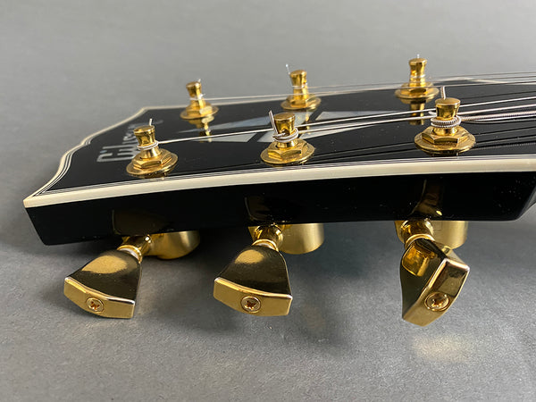 Close-up of black guitar headstock with gold tuning pegs and white binding on gray background