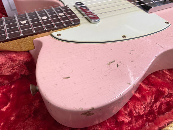 Close-up of pink relic electric guitar body with crackled finish and wear spots, rosewood fretboard, and white pickguard on red plush lining
