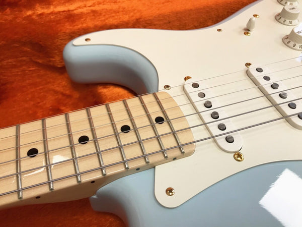 Close-up of light blue electric guitar neck and white pickguard with single-coil pickups on orange fabric background