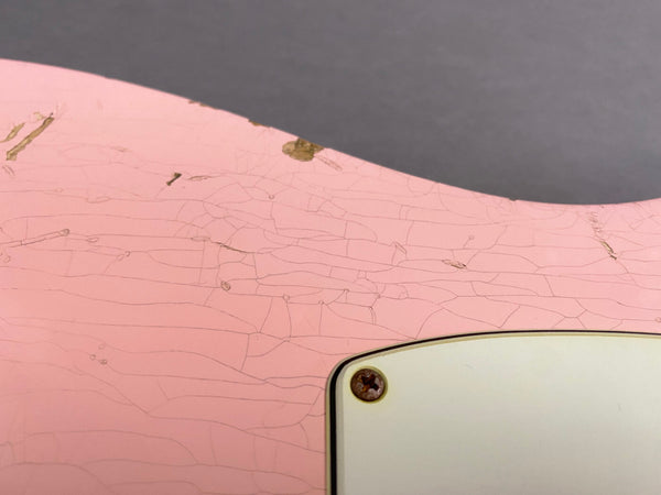 Close-up of pink electric guitar body showing vintage-style paint cracks and aging with screw detail on white backplate