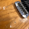 Close-up of natural wood electric guitar body showing chrome adjustable bridge with spring saddles and black pickup beneath strings, highlighting wood grain texture and finish imperfections.