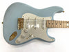 Light blue electric guitar with maple neck and fretboard, white pickguard, three single-coil pickups, gold hardware including bridge and output jack, volume and tone control knobs, close-up view on white background.