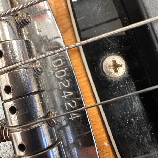 Close-up of guitar bridge and metal saddle with serial number 092424 visible on chrome plate, showing wound strings and black pickguard with screw.