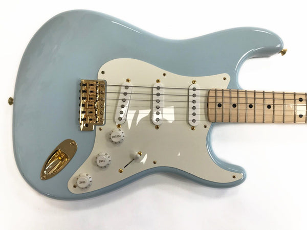 Light blue electric guitar with maple neck and fretboard, white pickguard, three single-coil pickups, gold hardware including bridge and output jack, volume and tone control knobs, close-up view on white background.