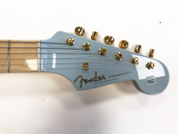 Close-up of light blue Fender Stratocaster guitar headstock with gold tuning pegs and maple fretboard, featuring "Fender" logo and "Stratocaster" text on white background