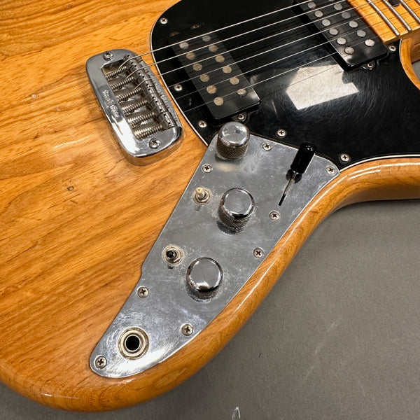 Close-up of natural wood electric guitar body showing chrome control plate with knobs, switches, and input jack, black pickups, and chrome bridge on gray background.