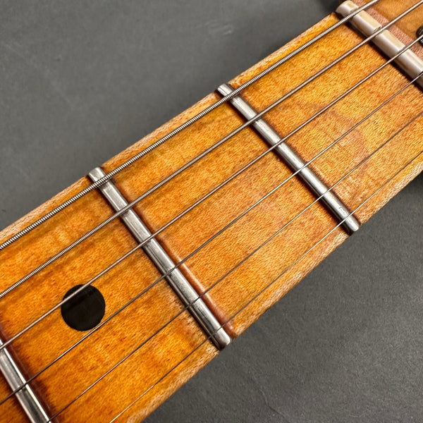 Close-up of guitar fretboard showing metal frets, wire strings, and black dot inlay on natural wood finish against dark background