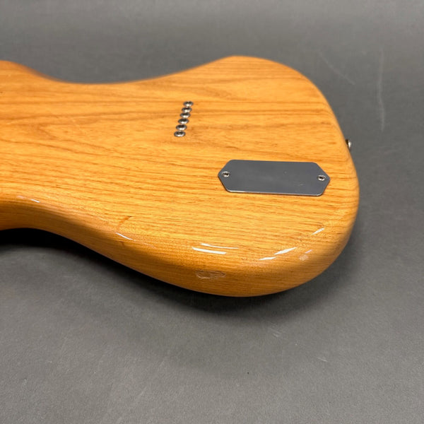 Close-up of the back body of a natural wood electric guitar with a metal control cavity cover and string ferrules on a gray surface