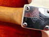 Close-up of guitar neck joint with metal neck plate engraved with serial number 197404 and stylized logo on white guitar body against red background