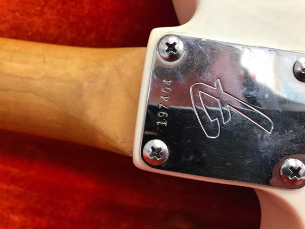 Close-up of guitar neck joint with metal neck plate engraved with serial number 197404 and stylized logo on white guitar body against red background