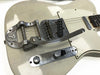 Close-up of vintage white electric guitar body showing chrome Bigsby vibrato tailpiece, single-coil pickups, and metal control plate with knobs and switch