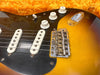 Close-up of electric guitar body showing sunburst finish, black pickguard, white single-coil pickups, volume and tone control knobs, and vintage-style bridge with six adjustable saddles