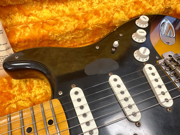 Close-up of vintage sunburst electric guitar body with black pickguard, three single-coil pickups, and white volume and tone control knobs on orange plush guitar case lining
