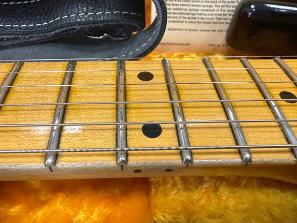 Close-up of maple guitar fretboard with silver frets and black dot inlays, six steel strings, and black leather strap on bright orange plush case lining