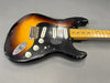 Vintage sunburst electric guitar with black pickguard and three single-coil pickups, maple neck with dot inlays, showing wear and paint chips on the body.