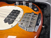Close-up of electric bass guitar bridge with "Music Man" engraved chrome bridge cover, four metal strings, black pickup, and amber wood body in guitar case interior