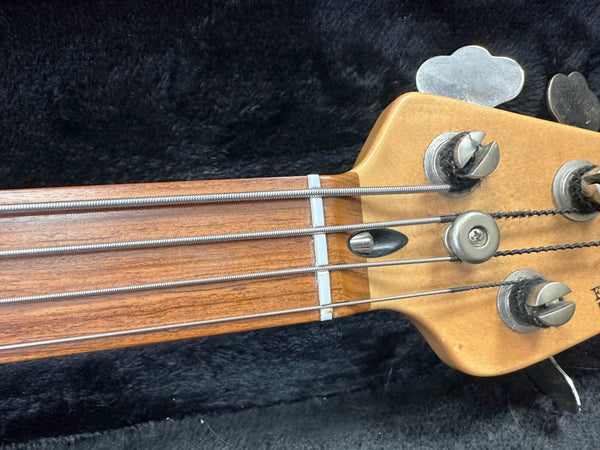 Close-up of electric bass guitar headstock and neck showing tuning pegs, strings, and nut on a dark fabric background