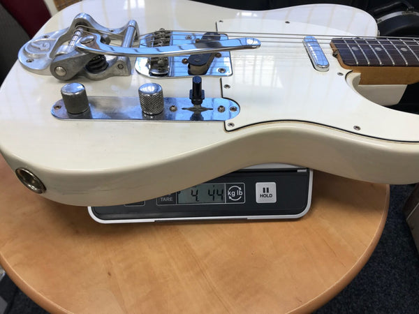 White electric guitar with Bigsby tremolo system and single-coil pickups on digital scale showing weight of 4.44 pounds, close-up of body and control knobs on wooden table