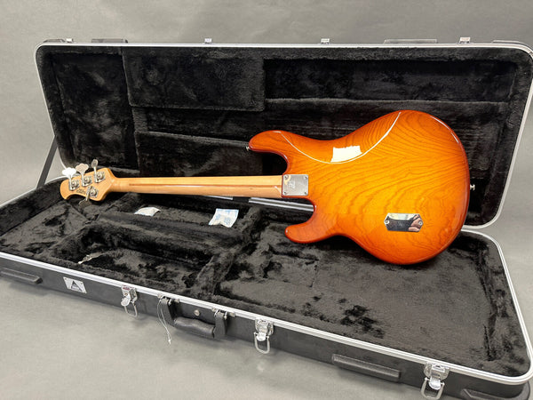 Electric bass guitar with sunburst finish lying in black plush-lined hard case, showing back side and maple neck with tuning pegs.