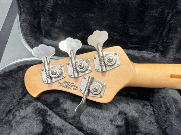 Back of bass guitar headstock with four tuning pegs, "Made in San Luis Obispo California USA" stamped on maple wood neck, resting in a black plush guitar case.