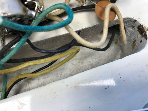 Close-up of vintage guitar wiring with cloth-covered wires in blue, yellow, black, and cream inside a white guitar cavity showing electronic components and soldered connections.