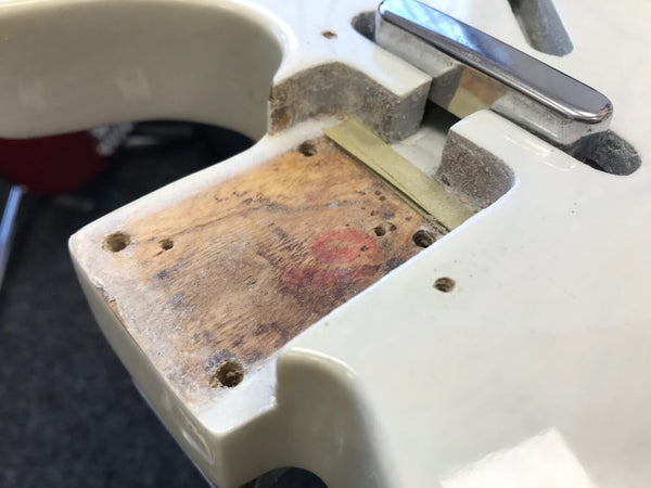 Close-up of white electric guitar body with neck pocket showing bare wood and mounting holes, chrome bridge saddle visible, part of disassembled guitar.