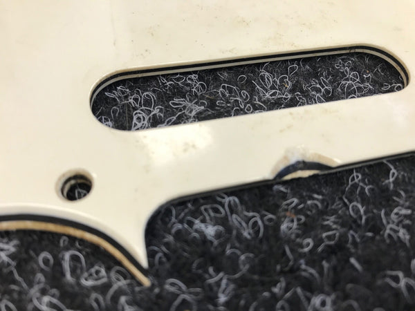 Close-up of white guitar pickguard with screw hole and black textured background showing wear and discoloration details