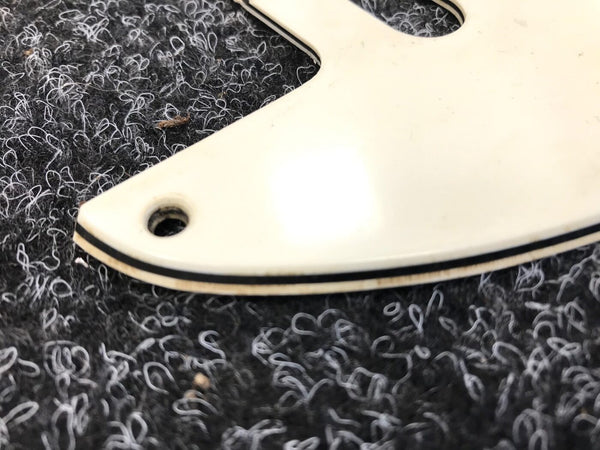 Close-up of white guitar pickguard corner with black and cream layered edging on textured black and white fabric background