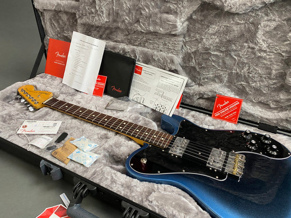 Fender electric guitar in metallic blue finish with rosewood fretboard, black pickguard, and chrome hardware, displayed inside a plush gray hardshell case with Fender manuals, setup instructions, warranty card, and accessories.