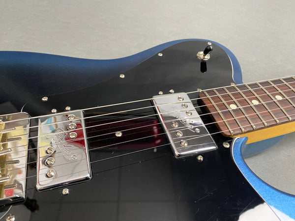 Close-up of blue electric guitar body showing black pickguard, chrome Fender pickups, switch, and rosewood fretboard with dot inlays