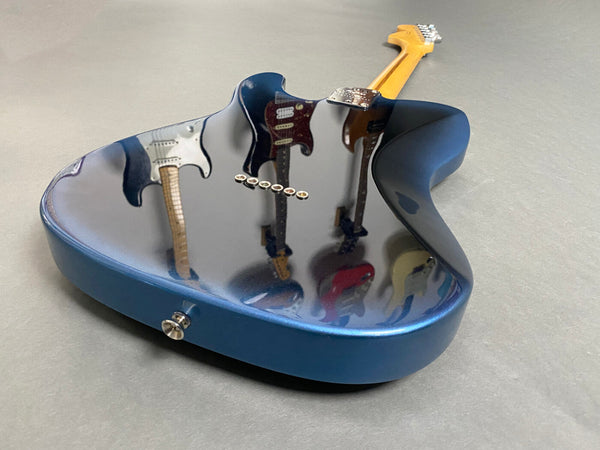 Blue electric guitar body with high-gloss finish reflecting multiple guitars on gray surface.