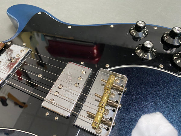Close-up of blue Fender electric guitar body showing pickups, bridge with brass saddles, volume and tone control knobs, and strings
